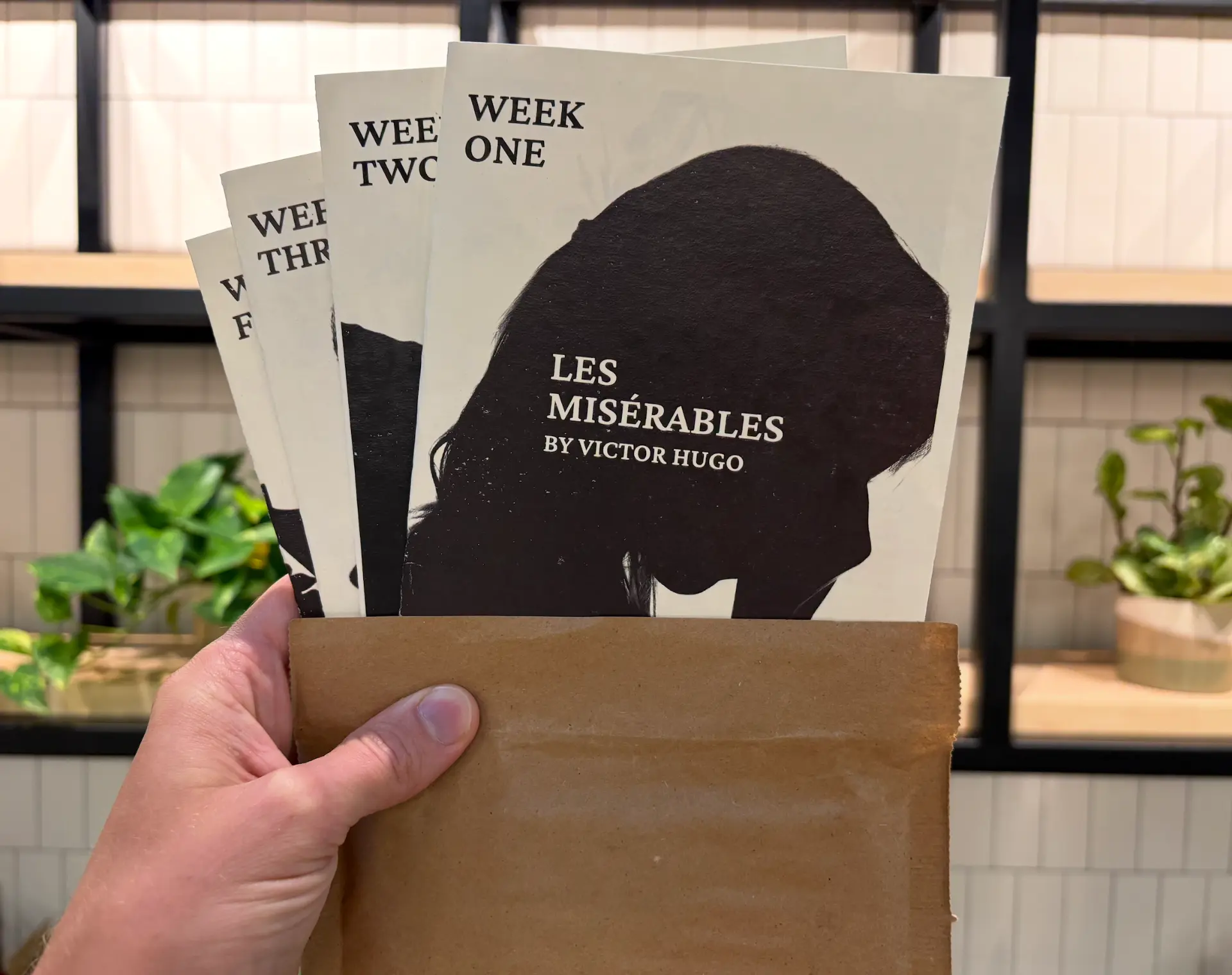 A hand holding a Human by Hand mailer with four weekly packets visible, including Week One: Les Misérables by Victor Hugo