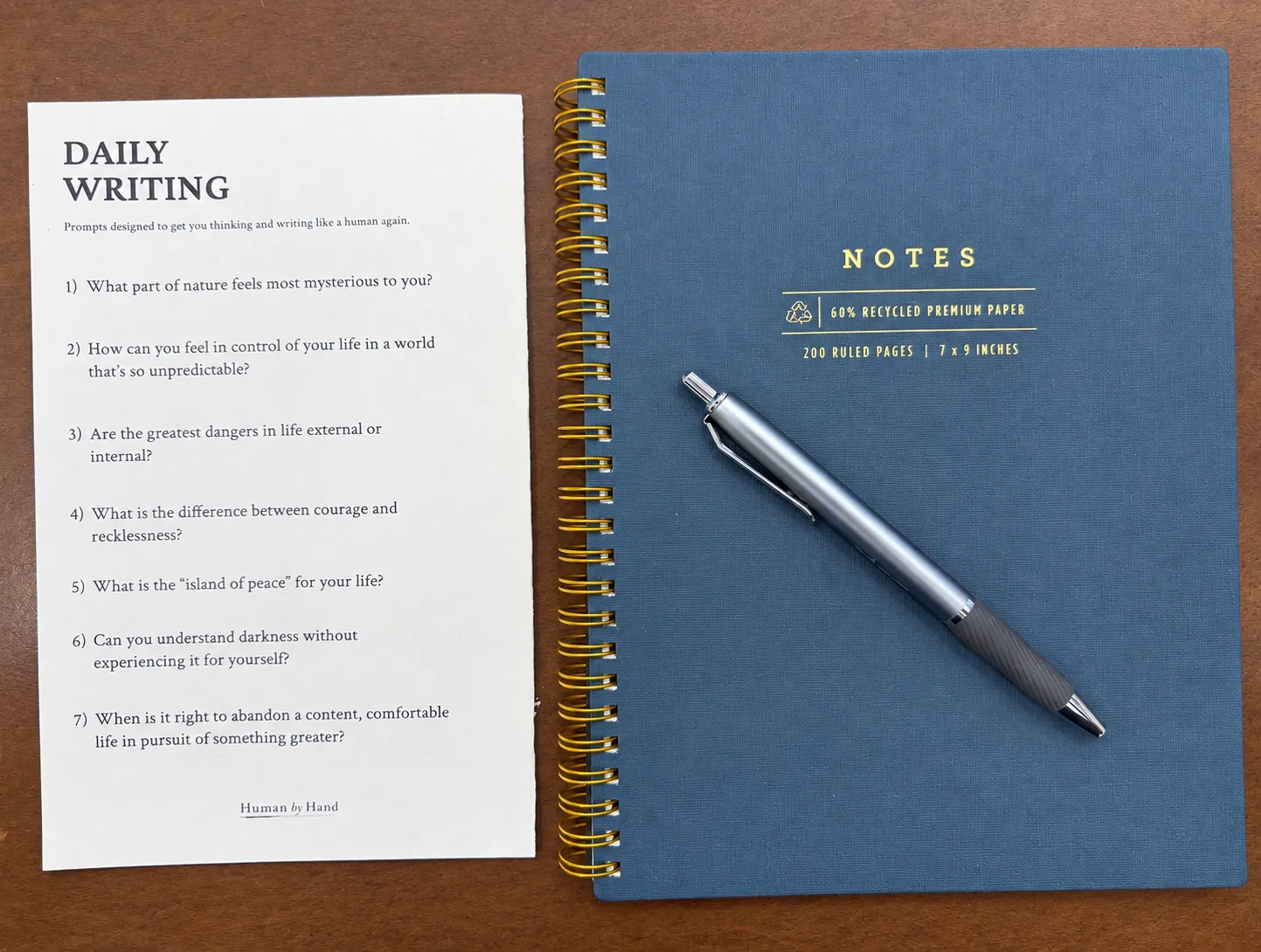 A Daily Writing prompt sheet beside a blue spiral notebook and pen, showing seven reflection questions